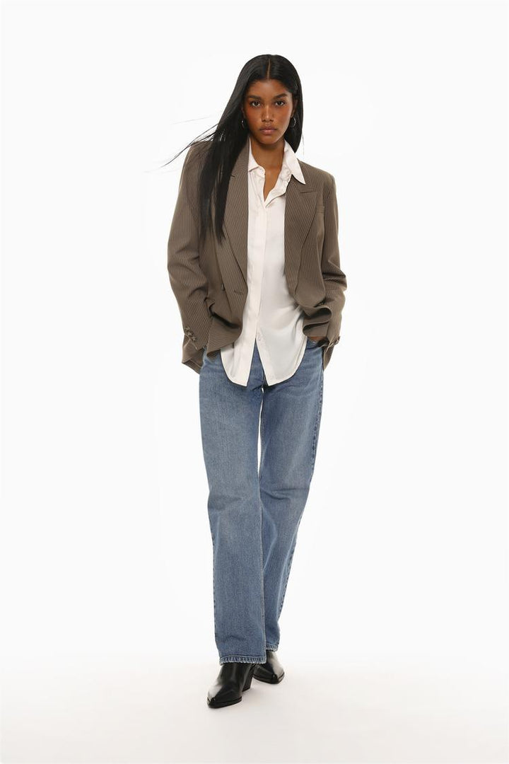 A full-body view of a model wearing a white long-sleeve satin shirt. The image showcases the shirt's elegant drape, glossy finish, and relaxed yet sophisticated fit. Perfectly paired with neutral-toned trousers for a polished look.